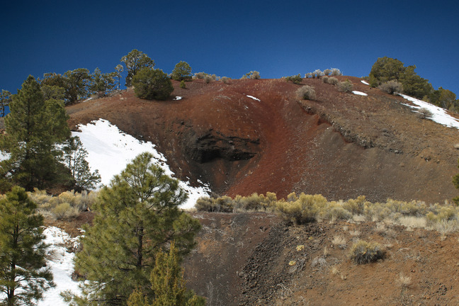 An upside-down cone of rust, tan, and black lava cinders reaches into a cloudless sky.  Patches of snow, trees, and shrubs dot the surface.