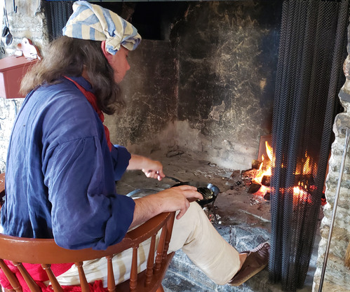 A person in historic clothing stirs food cooking in a skillet over coals in a stone hearth with a fire burning.