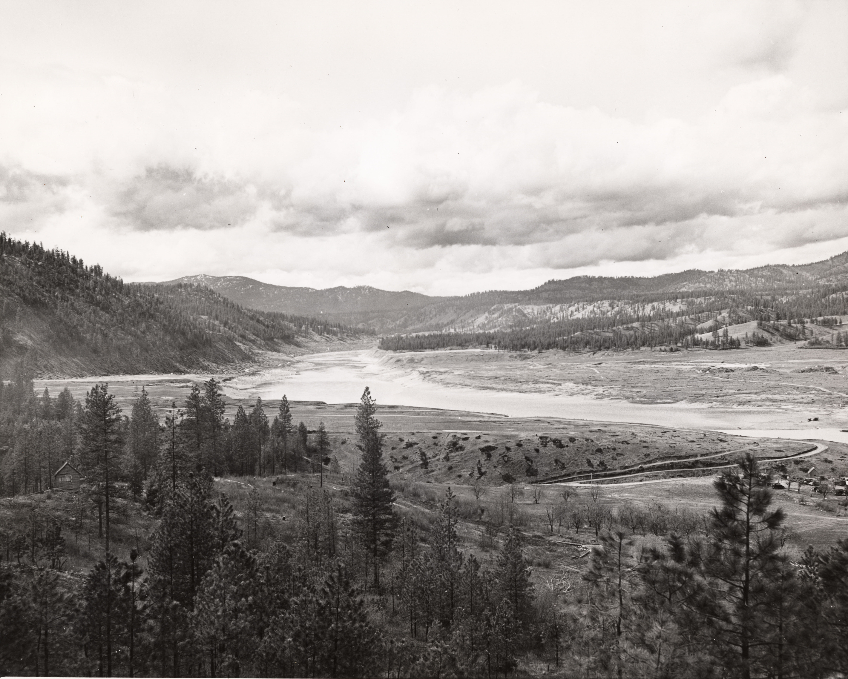 Black and white photograph of a river in a wide valley with cleared flats on either side
