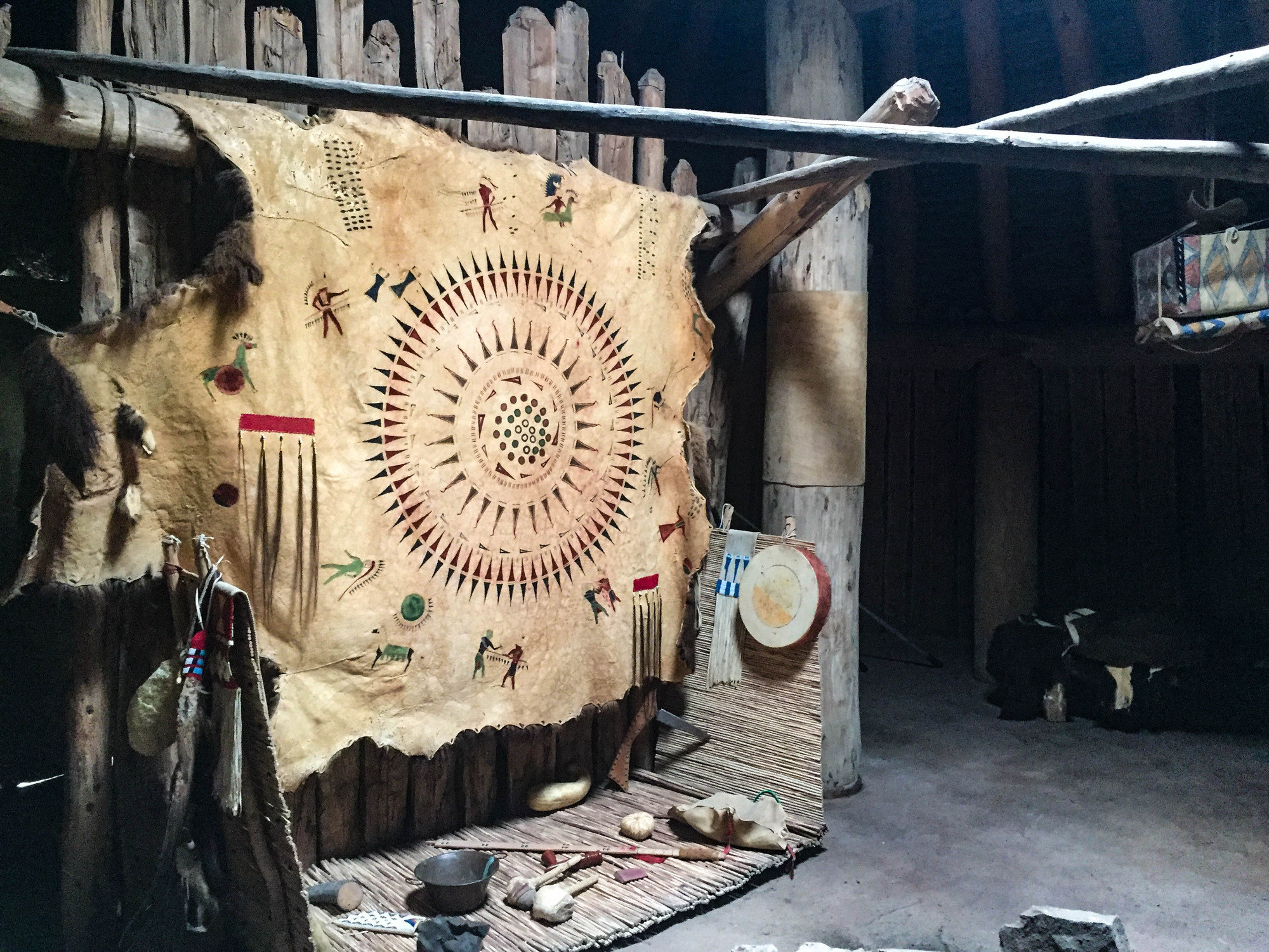 Various Native American items on a mat underneath a hanging animal hide decorated with paintings