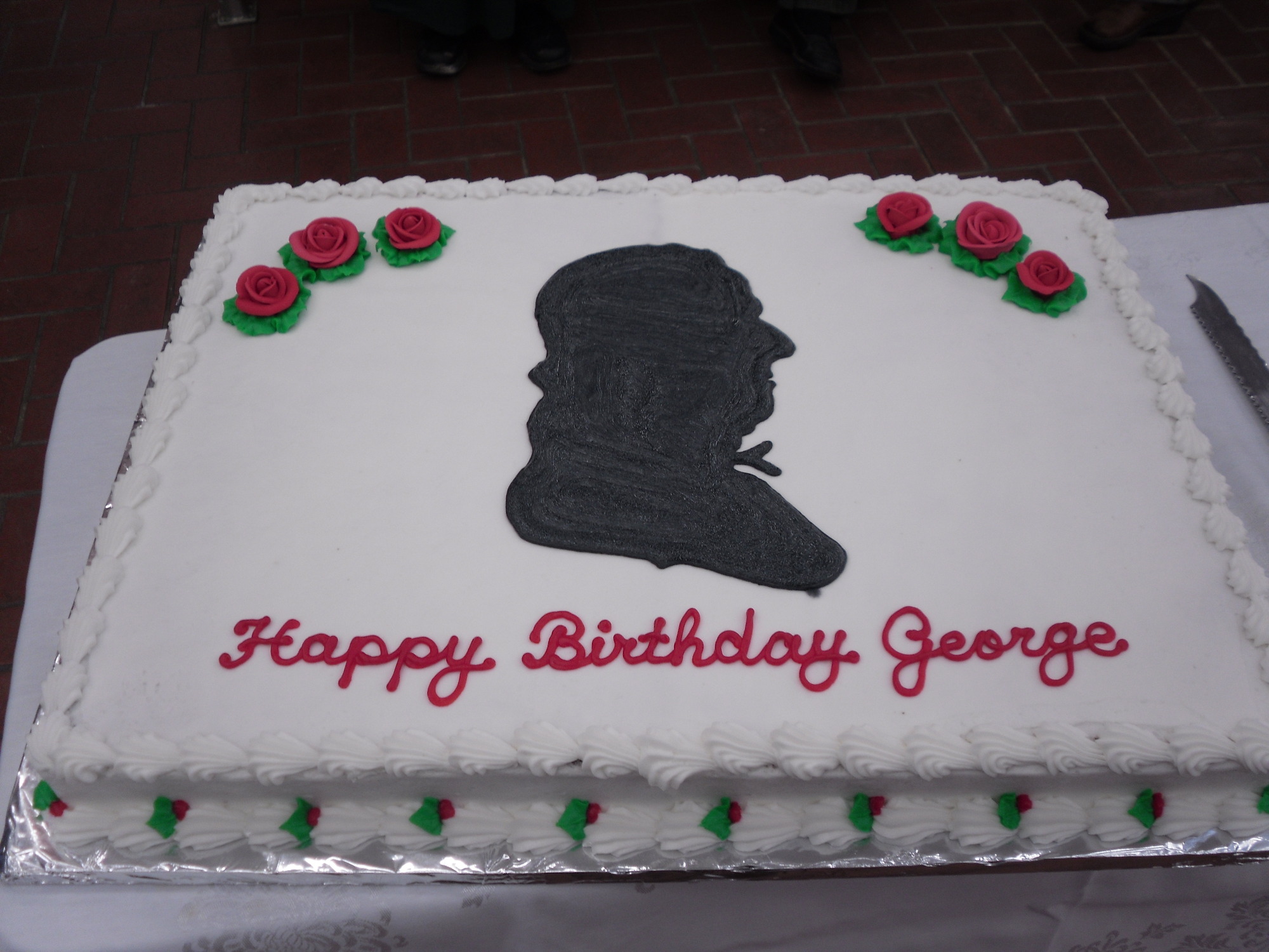 White icing cake with red roses on it and Happy Birthday George written red with a black silhouette of George