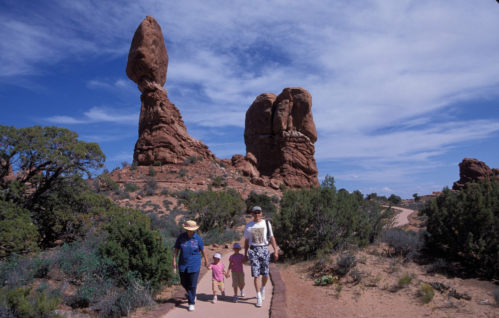 two adults and two children walk on a paved trail in front of a tall, balanced rock.