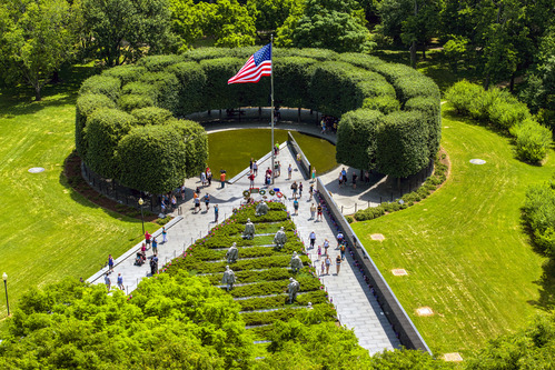 An aerial view of the Korean War Veterans Memorial: A 'v' shaped walkway leads towards a pool of remembrance; inside the 'v' are statues of soldiers representing different parts of the armed forces. A marble wall with the faces of veterans lines the right side of the 'v'. 