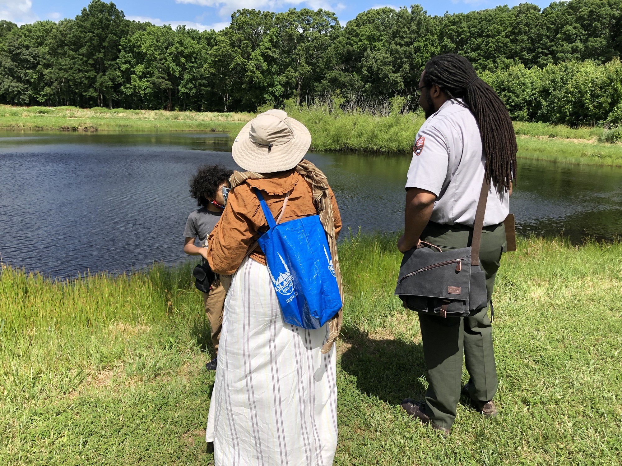 A park ranger, a child, and a woman can be seen standing on grass in the foreground. Before them is a pond with tall grasses at the far right side of the pond. Tall trees can be seen in the background.