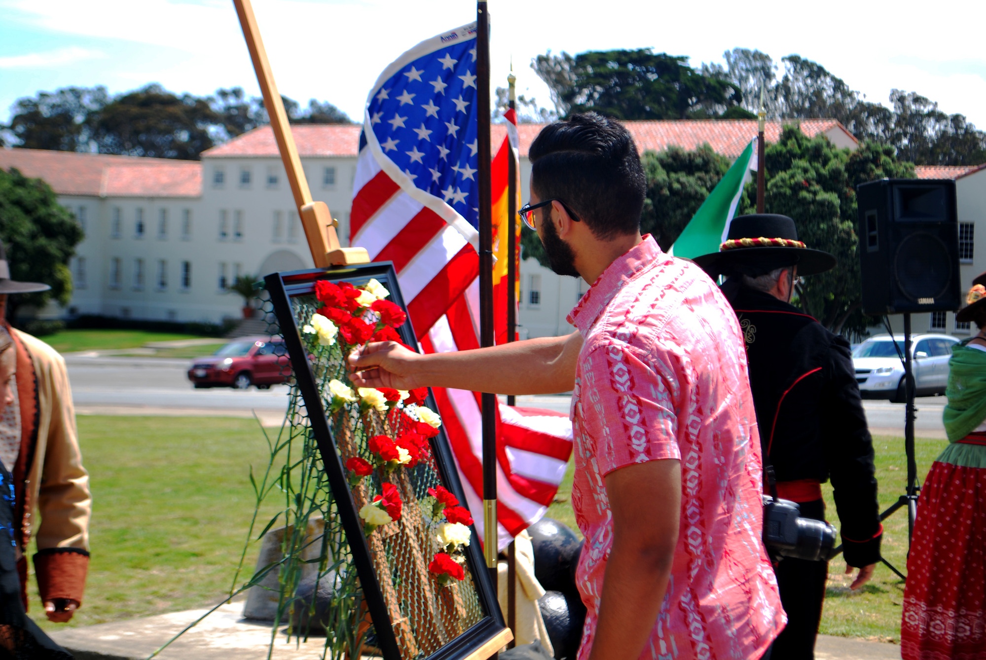 A person wearing a pink and white button-down short-sleeved shirt puts a carnation into a frame at a ceremony on a lawn