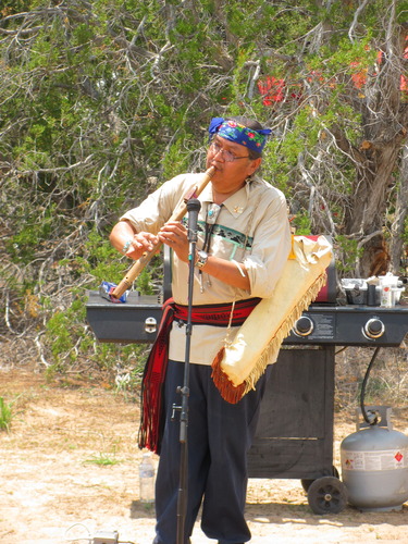 A man with a blue floral headband and leather case on his side plays a wooden flute near a microphone.