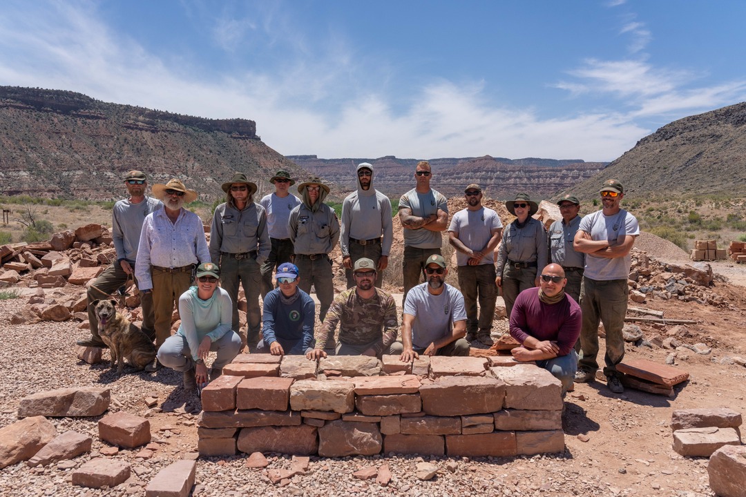 A group of people and a dog standing and crouching down for a photo, with a sandstone brick wall in front of them and red and green sandstone canyons behind them. 