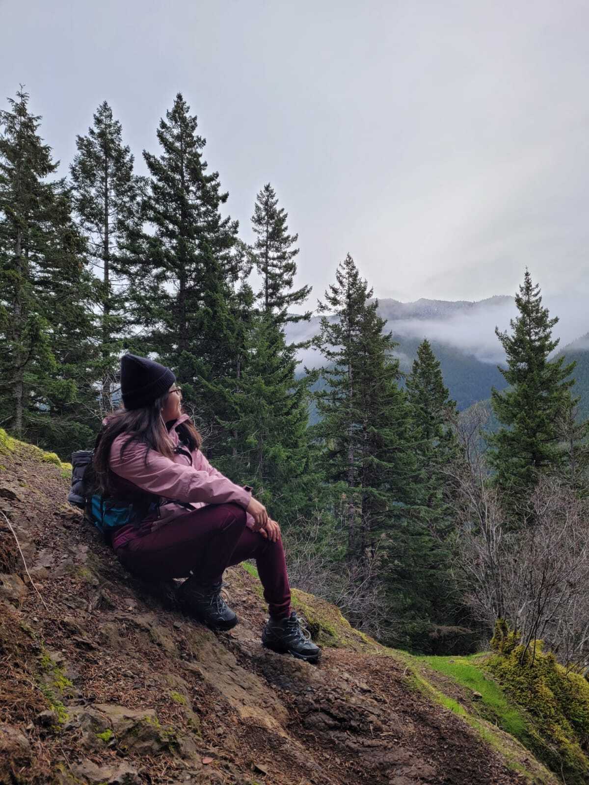 Woman sitting down on side of hill surrounded by trees, while admiring the view ahead.