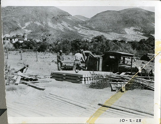 This is an historic black and white photograph from the Scotty's Castle Historic Photograph Collection, Death Valley National Park of three unidentified employees moving concrete fence posts onto truck with Scotty's Castle Main House in distance. October 2, 1928. Photographed by Mat Roy Thompson.