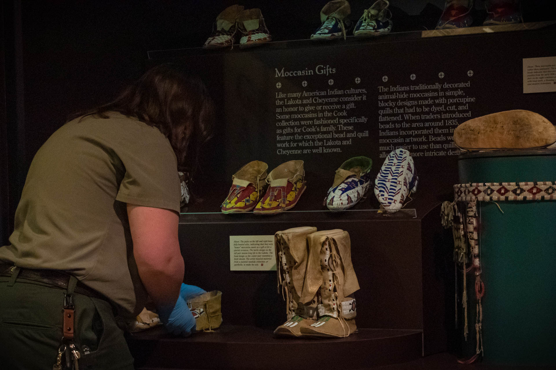 Female uniformed ranger with blue gloves lifts moccasins out of open museum display case.