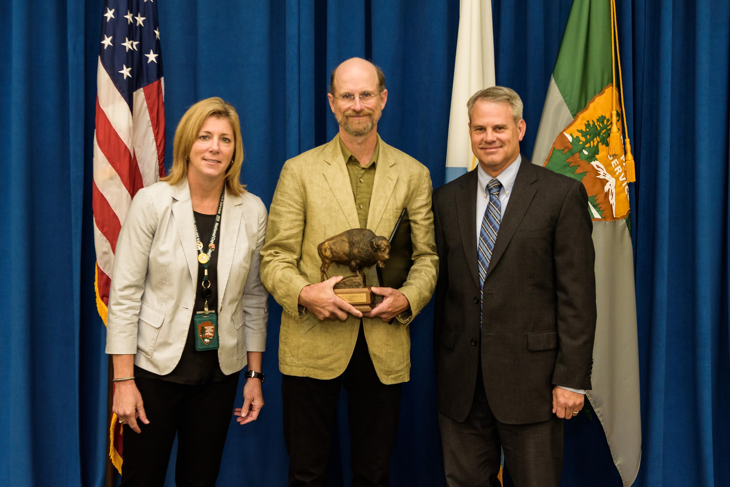 a man in a tan suit holds a bison statue and stands between a man and a woman in business suits. The background is a blue curtain, an American flag, and a National Park Service flag.