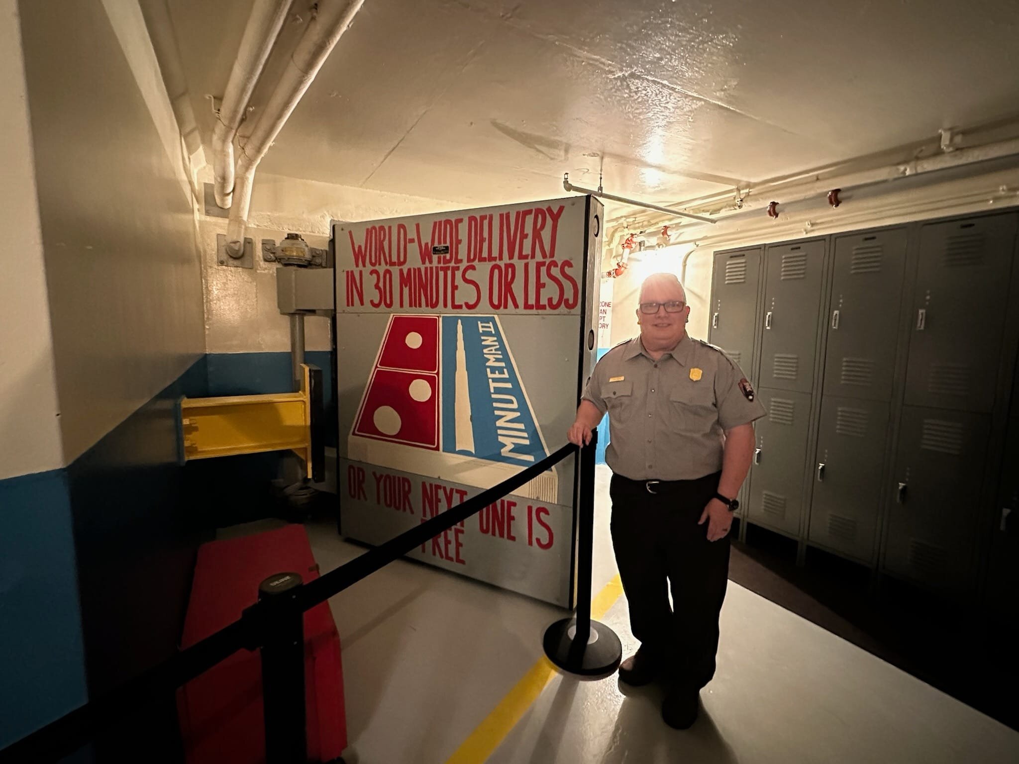 Man in a park ranger uniform of gray shirt and green pants stands next to a large square gray door painted with a domino's pizza logo in red, white, and blue. Words in red "world wide delivery in 30 minutes or less or your next one is free"