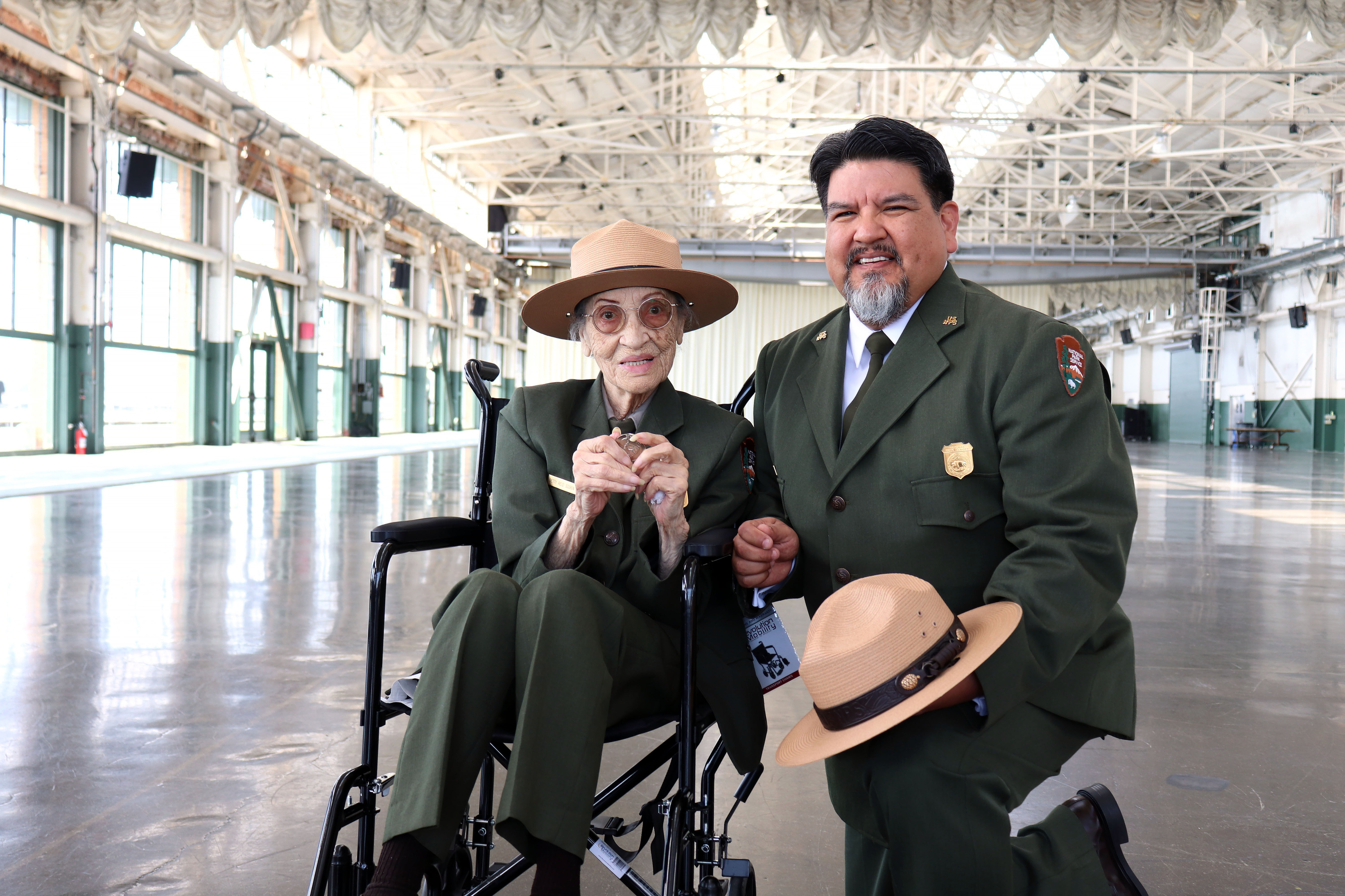 A senior African American woman in a ranger uniform sits in a wheelchair and is posing with an older man in a ranger uniform. He is of Native American decent. 