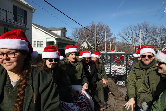 Park Rangers in Santa hats sit facing each other and the camera on a wooden trailer. 