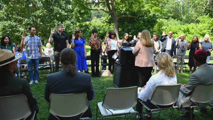 More than a dozen people stand with right hands raised in front of a woman standing at a podium.