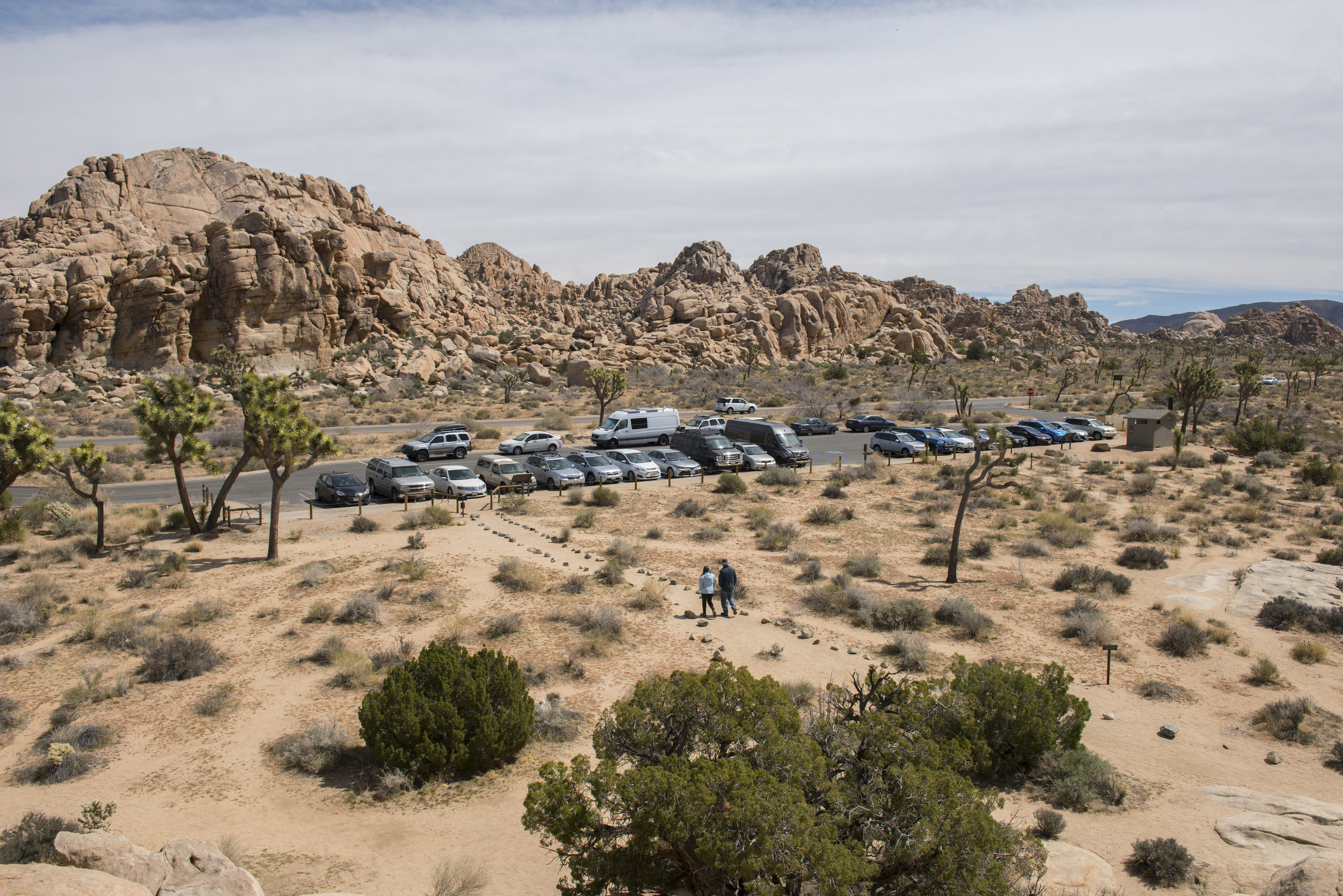 A small parking lot filled with vehicles surrounded by a desert landscape with some rocky formations in the background. Two people walk on a trail heading towards the parking lot. 