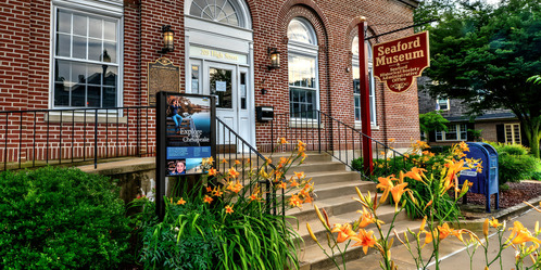 Entrance of the Seaford Museum with orange lilies, brick walls, and a red sign. Transcribed Text: Signboard: "Explore Chesapeake" • Red sign: "Seaford Museum Seaford Historical Society Admin Office" • Above the door: "203 High Street"
Image