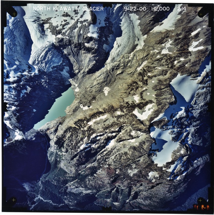 Color aerial photograph of glacier, snowfields, and alpine lake on mountain peak.