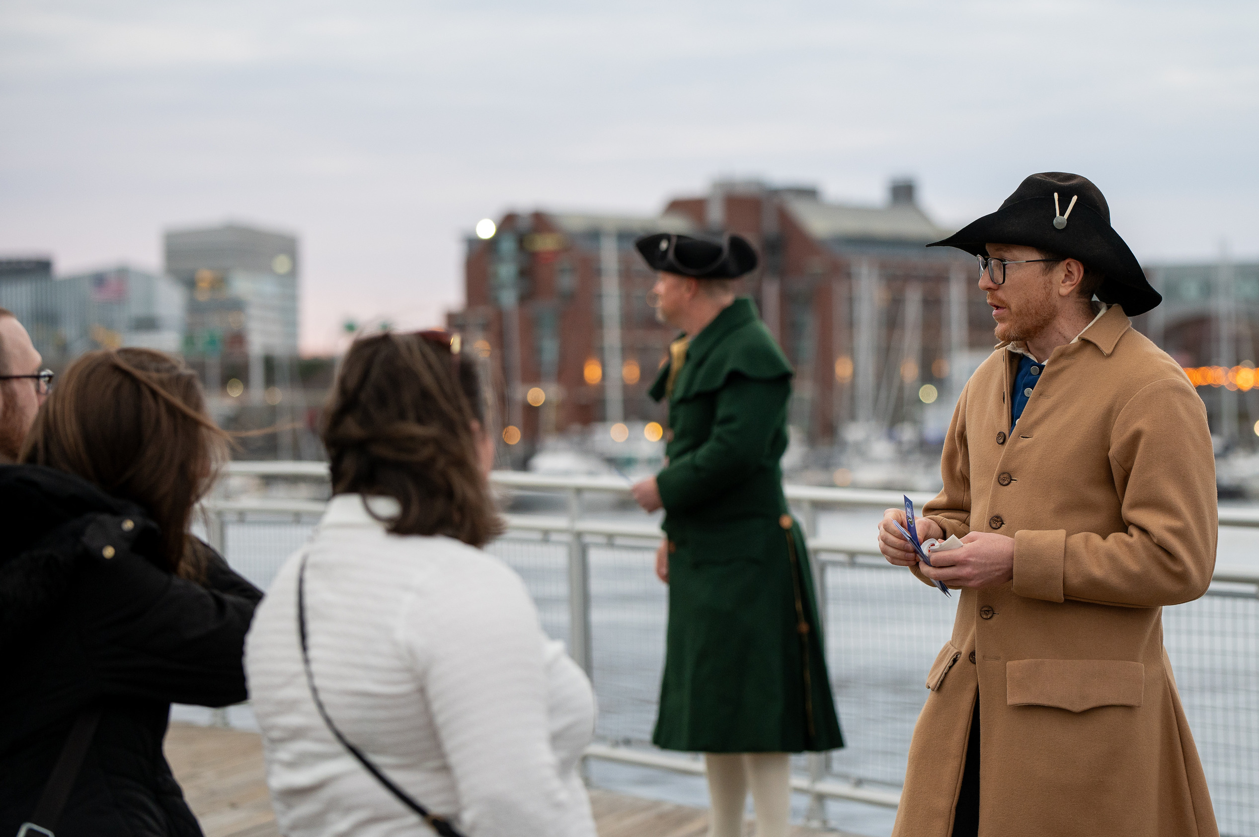 Two men in colonial era kit talking with event attendees. The harbor with boats are in the background. 