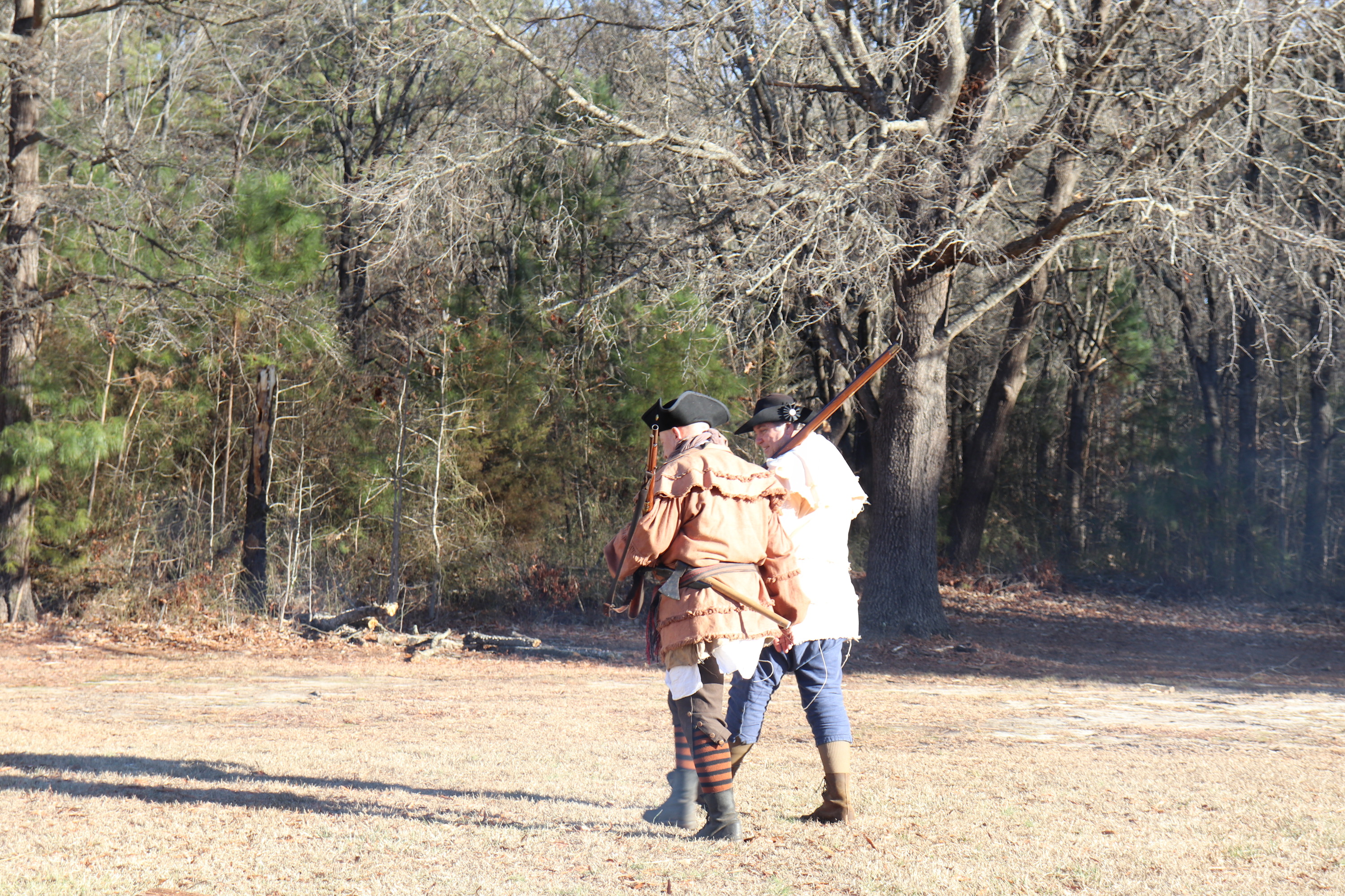 Two living historians dressed as militia return to camp with muskets shouldered. woods lie just in the background as they make their way through a field.