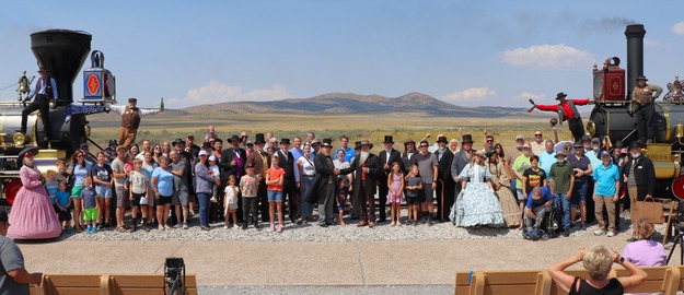 People standing together in front of two colorful historic locomotives.  Two Locomotives perch hanging over the front of locomotives with bottles held high. 