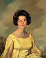 Head and torso portrait of a woman with a slightly curled brown hair, shiny earrings, and a delicate yellow dress, with a backdrop of a monument 
