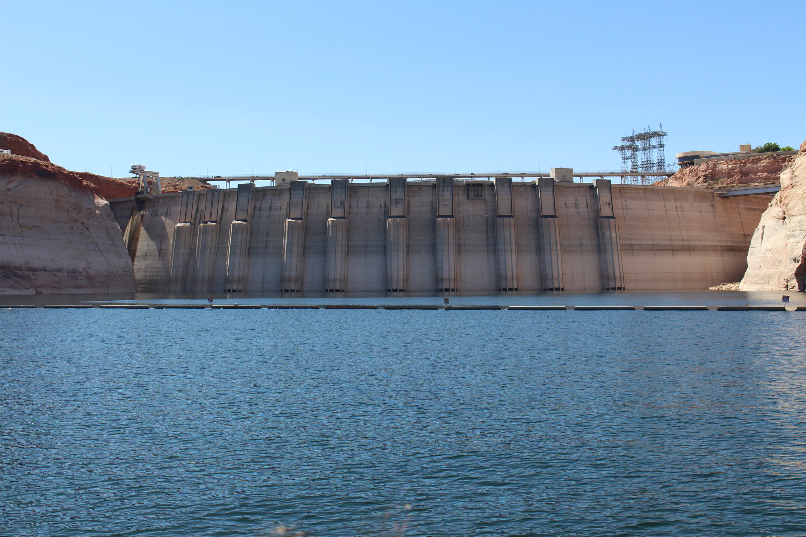Concrete structure holding back water in steep rock canyon. A metal suspension bridge with vehicles rises above the top of the structure. 
