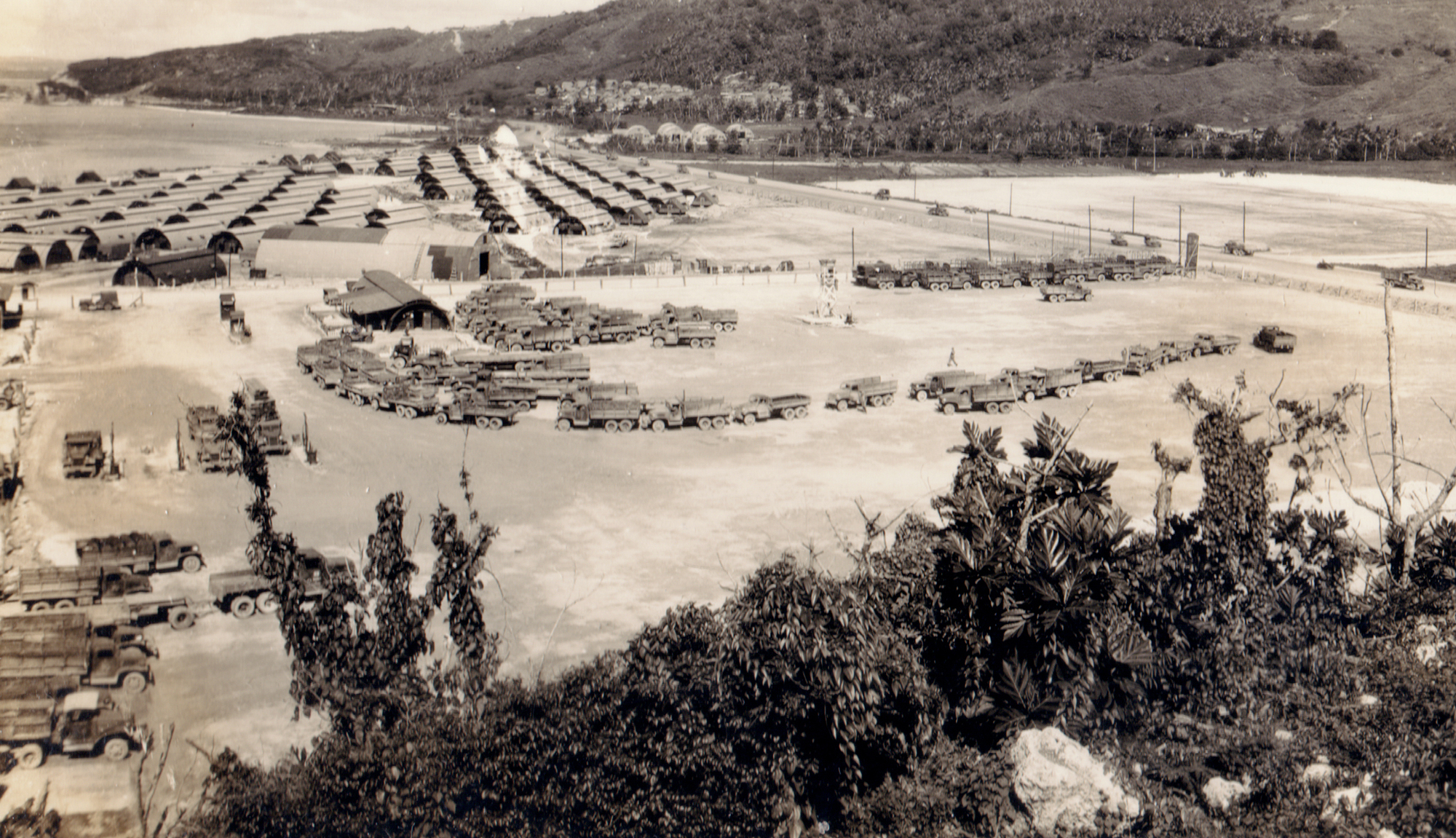 Black and white photo of a line of trucks next to a series of Quonset huts.
