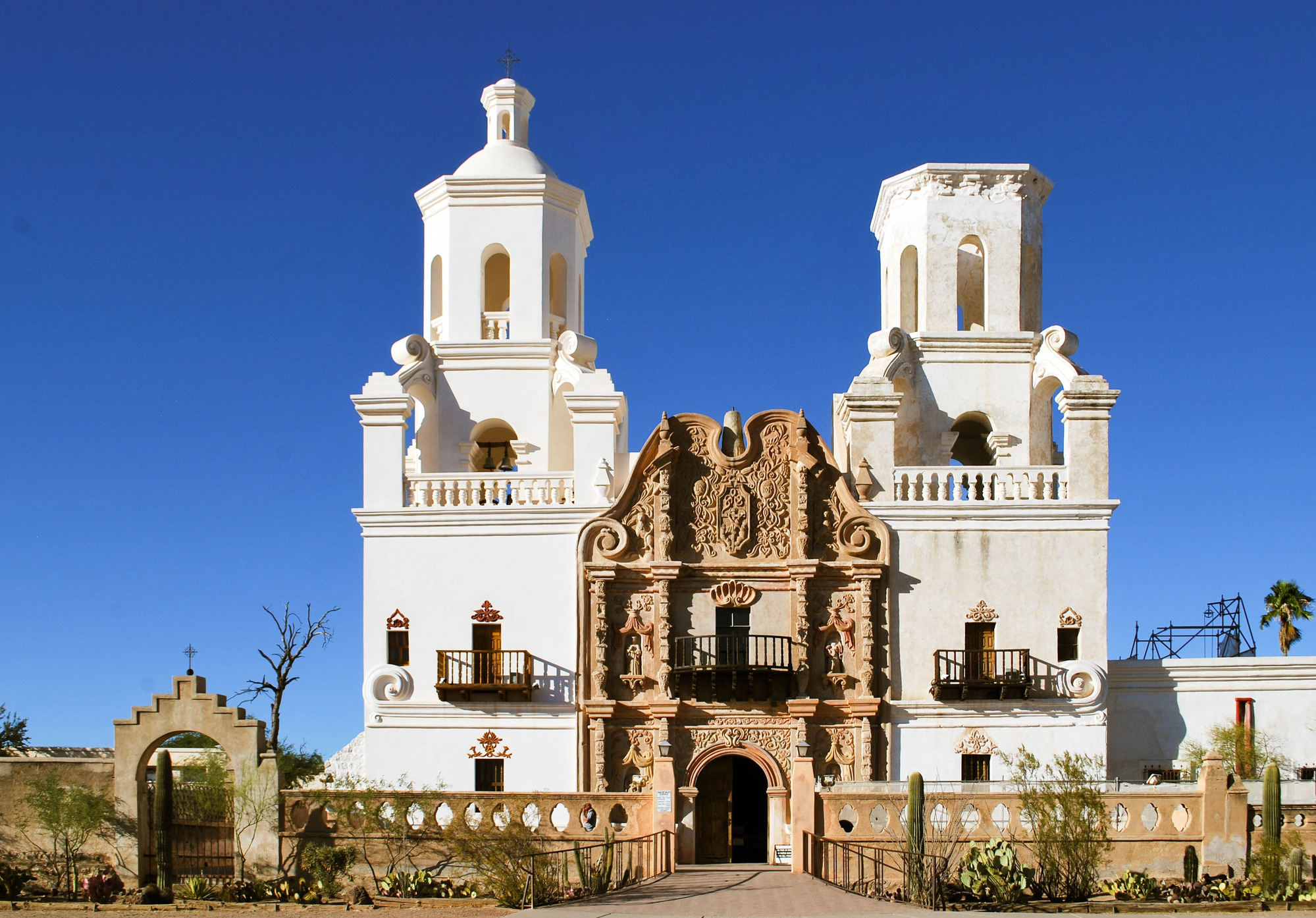 A long stone wall with an archway sits in front of a white mission with two bell towers on each side with an intricately decorated brown stucco facade in the center