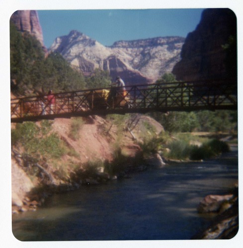 Man driving sweeper equipment across the new Grotto footbridge after its placement on stone abutments. Men working on trail in background.
