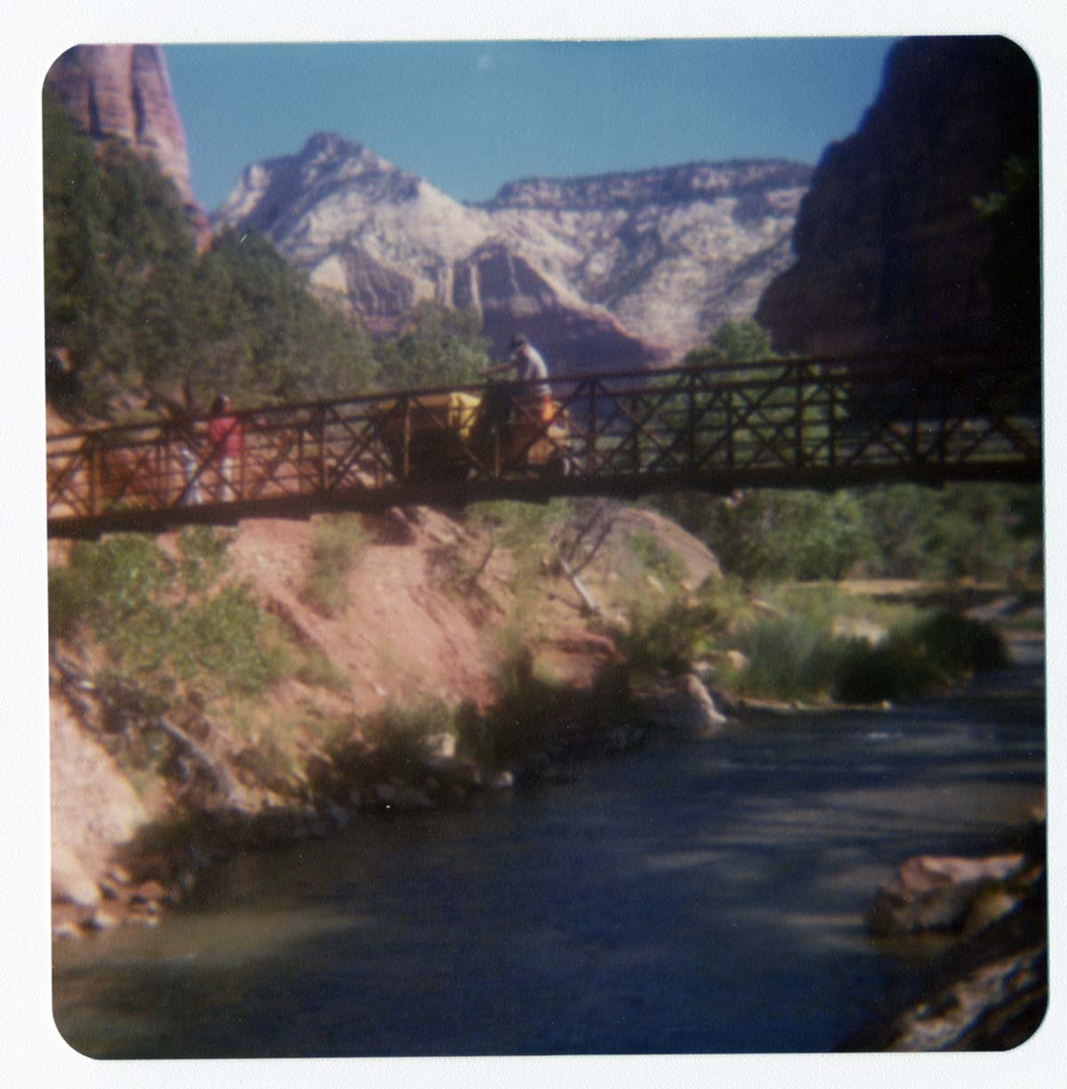Man driving sweeper equipment across the new Grotto footbridge after its placement on stone abutments. Men working on trail in background.