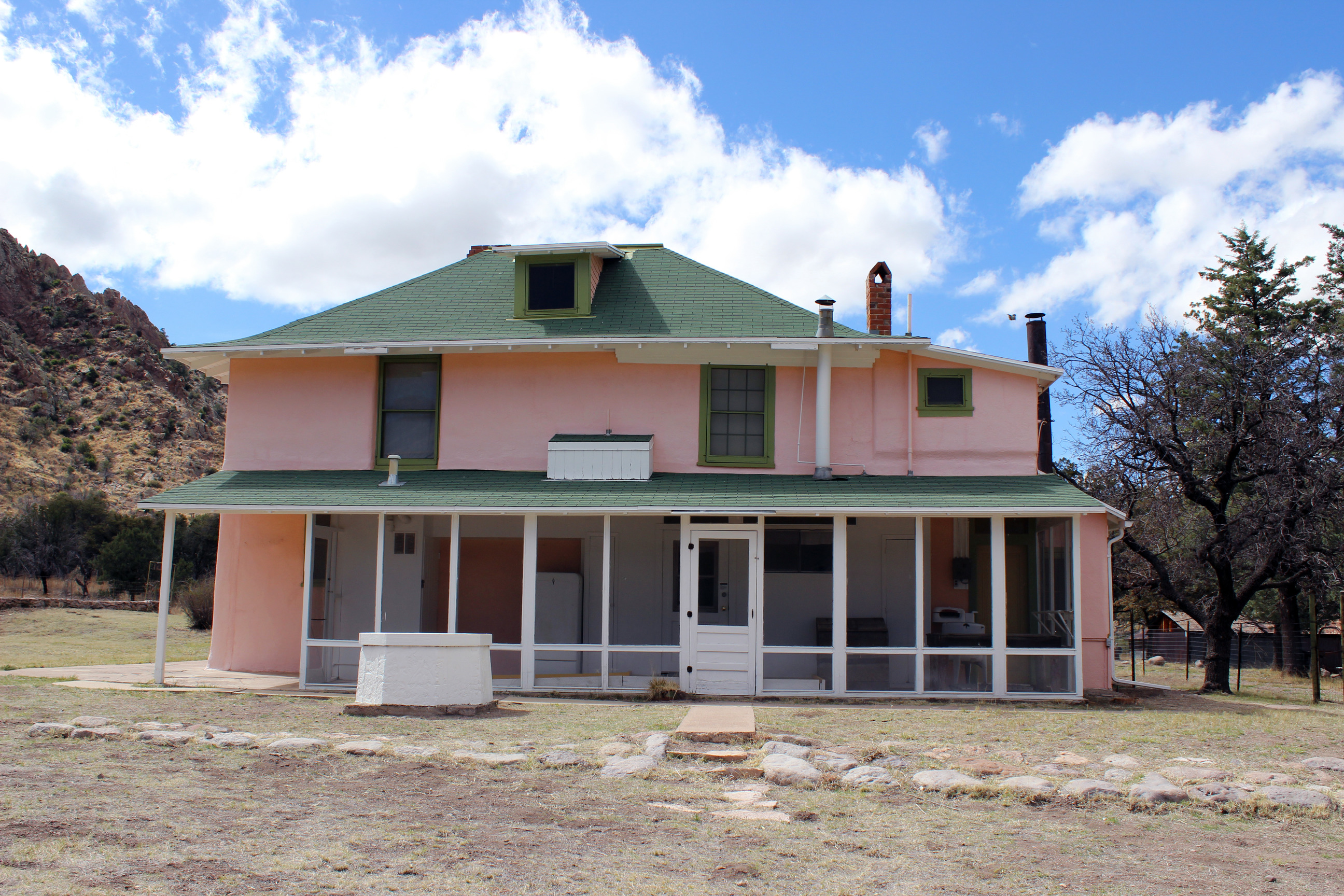 Pink adobe hosue with green roof and white porch and white well. 