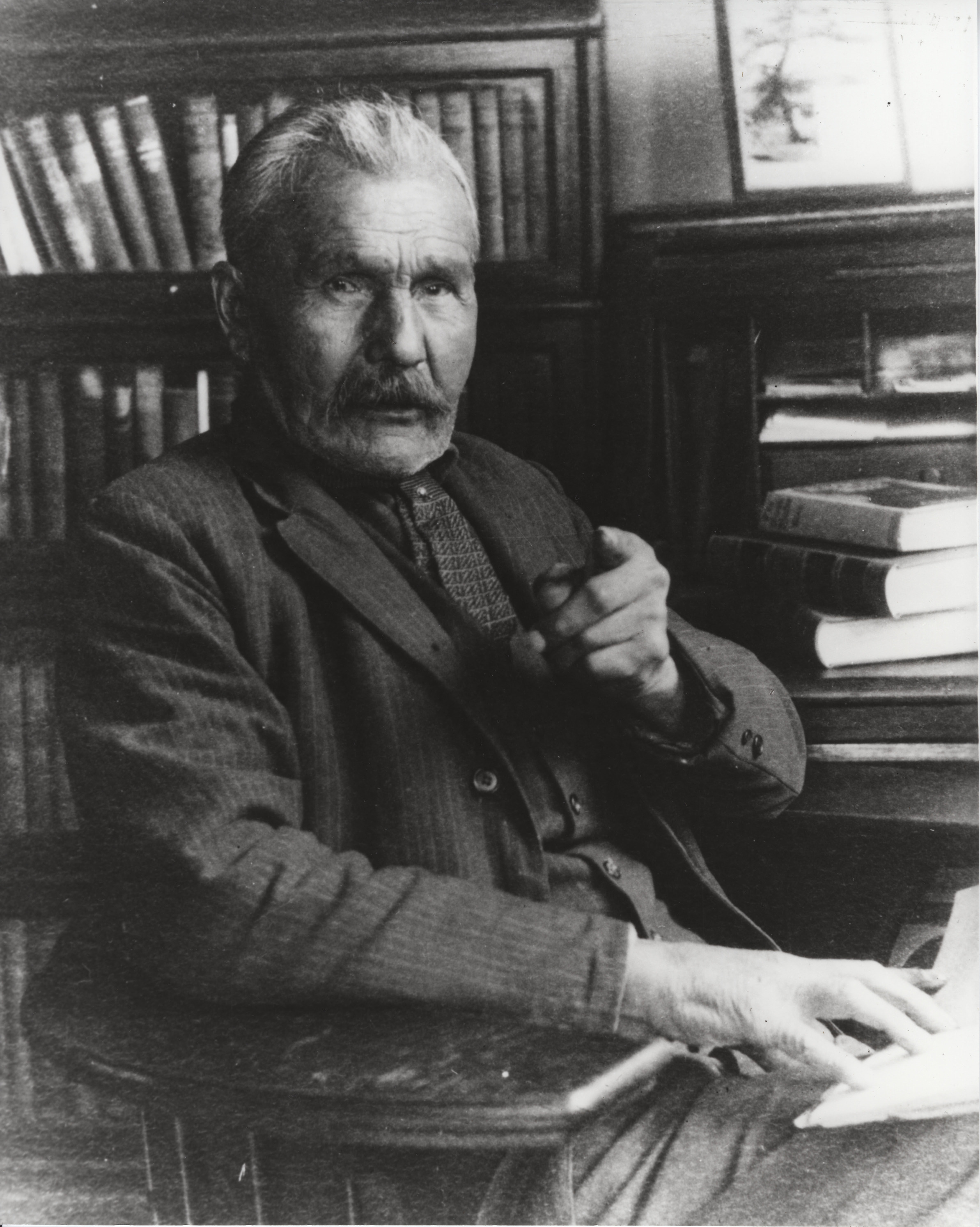 A black and white photograph of an older man sitting for a portrait in a study, gesturing at the camera