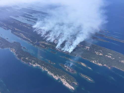 Aerial view of fire burning on island in a lake. Smoke lifts from the forest and fills the sky. 
