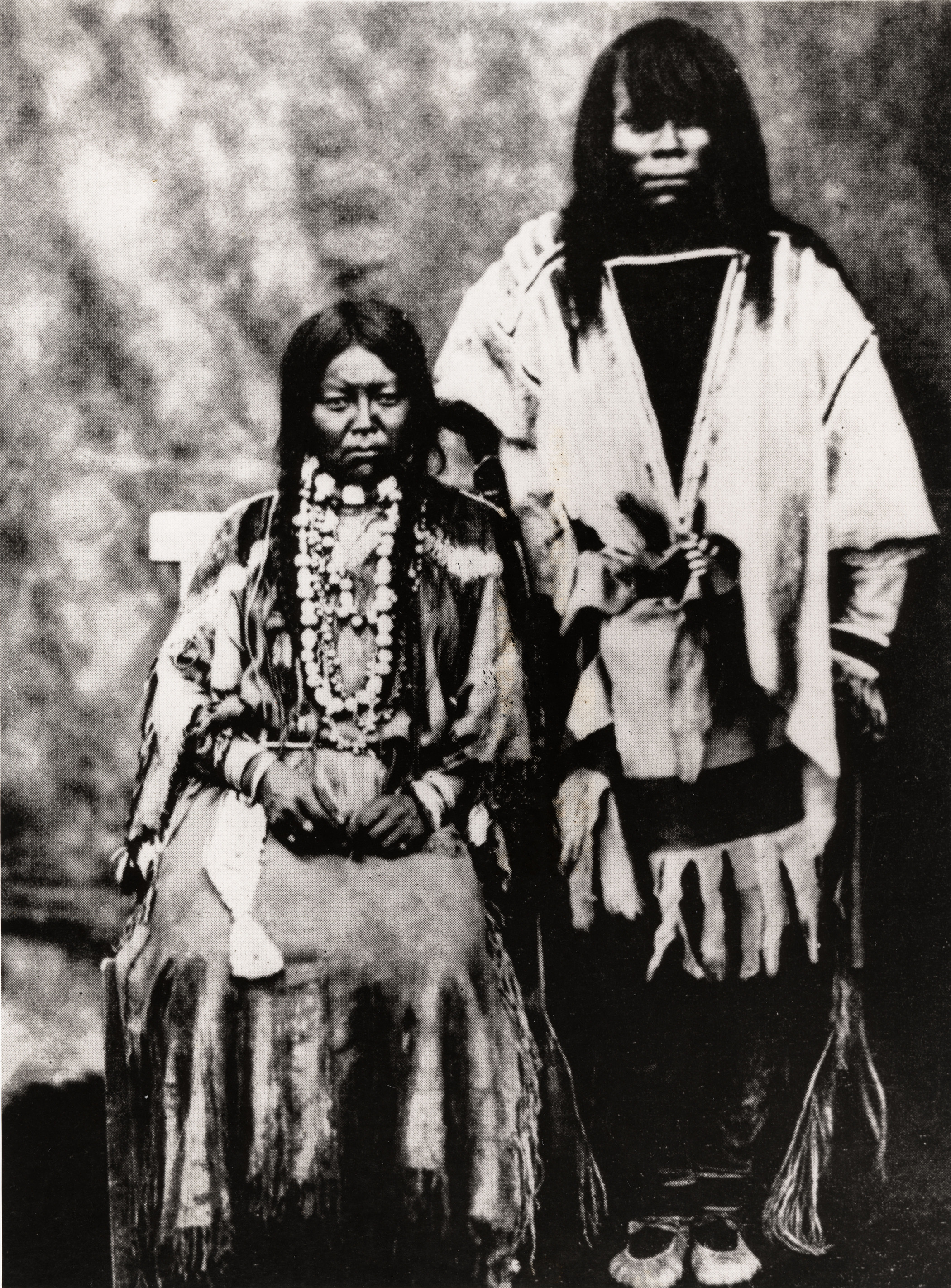 Black and white photograph of two American Indians posing for a portraits, one standing and one seated