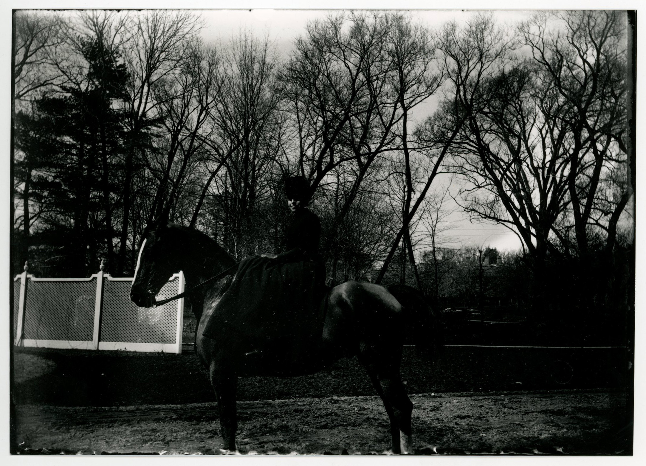 A white woman in riding hat and habit sits sidesaddle on a dark-colored horse. White fence in left midground. Trees in background.