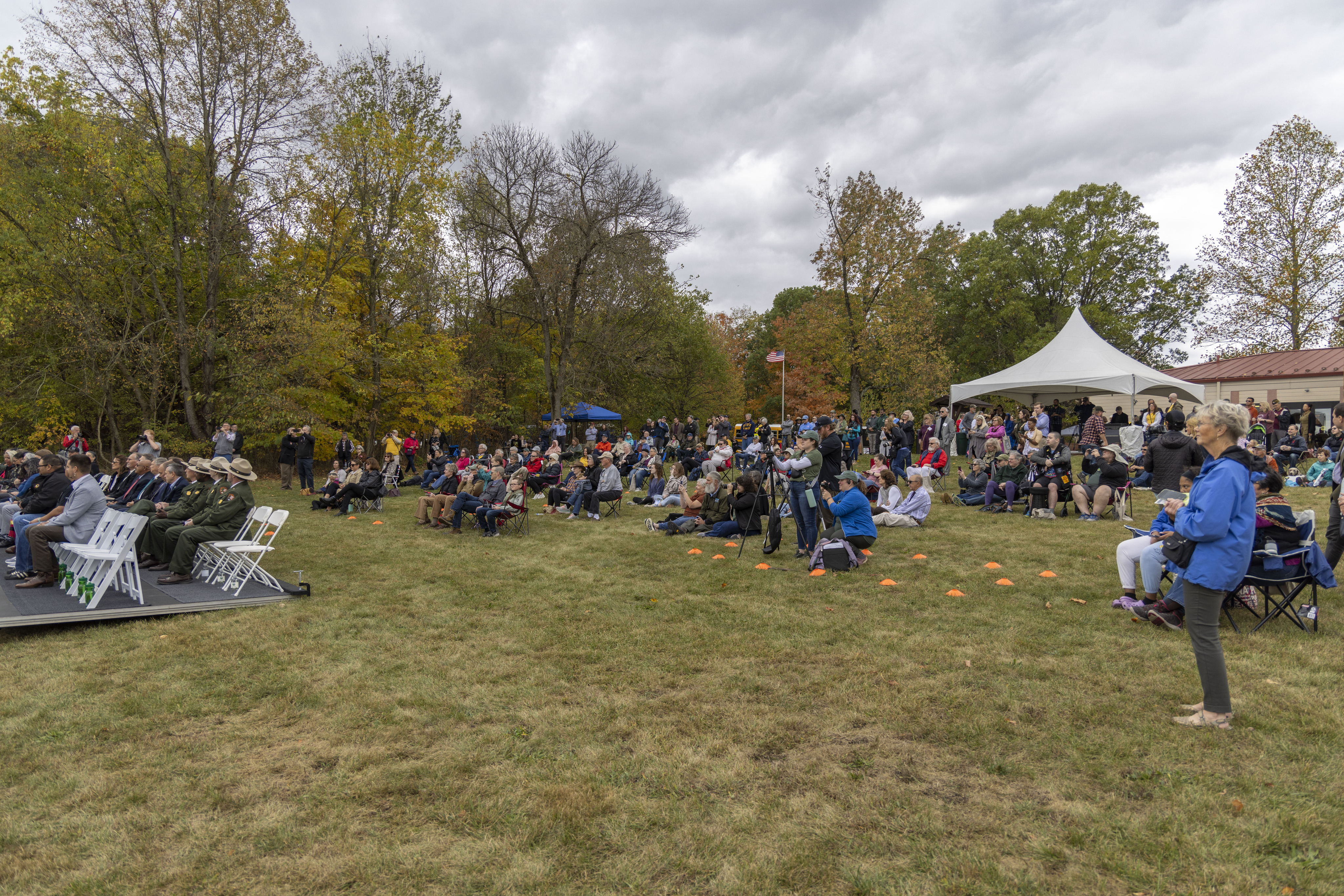 A green grassy field with several people in warm clothing and jackets stand and sit while listening to other people speak to them on a stage that is out of picture to the left. Some people in the crowd are seated in while chairs at the left side of the image.