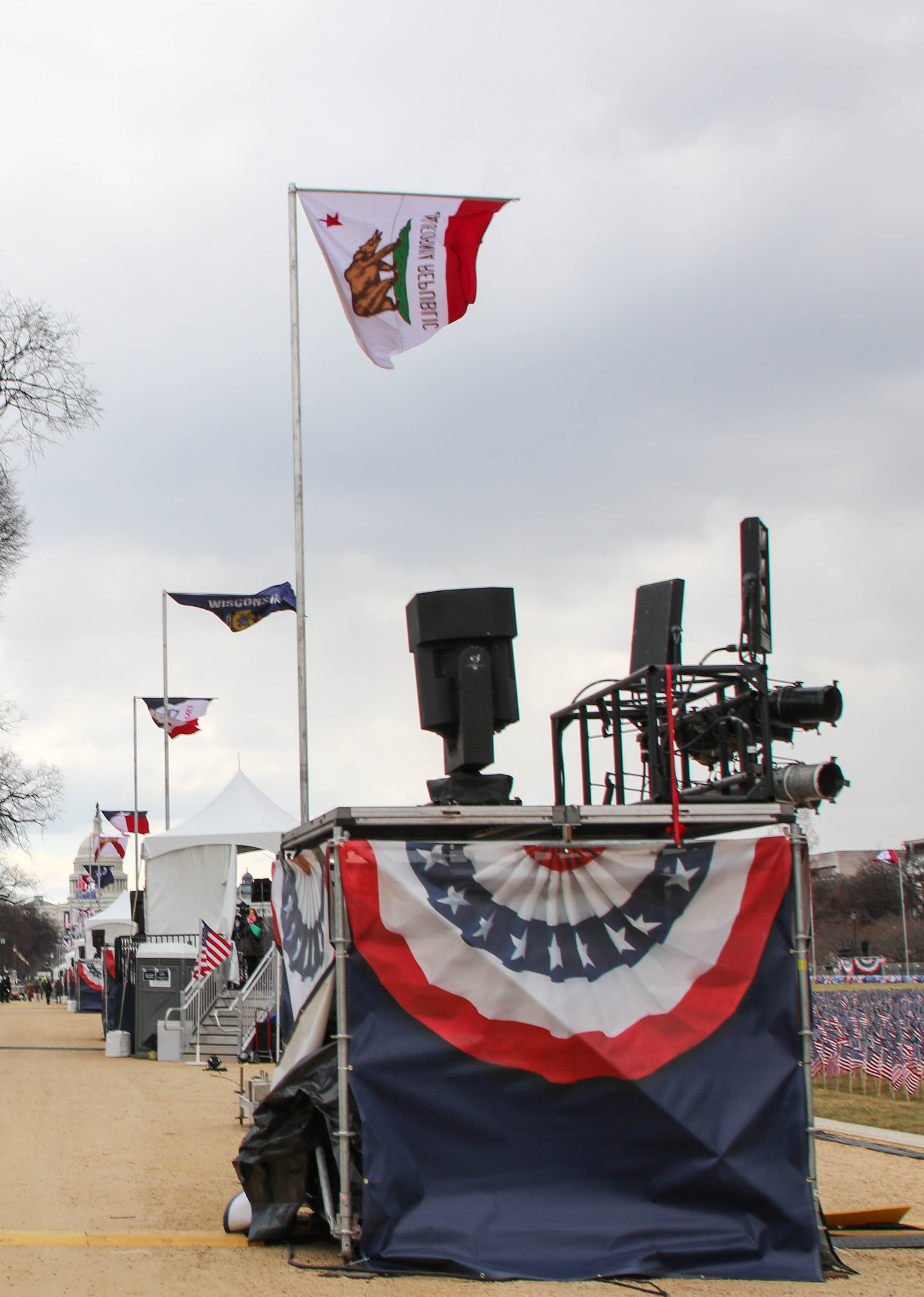 A wide pathway lined with spaced lighting stands on platforms and state flags on flagpoles. A lawn decorated with small U.S. flags is next to the pathway.