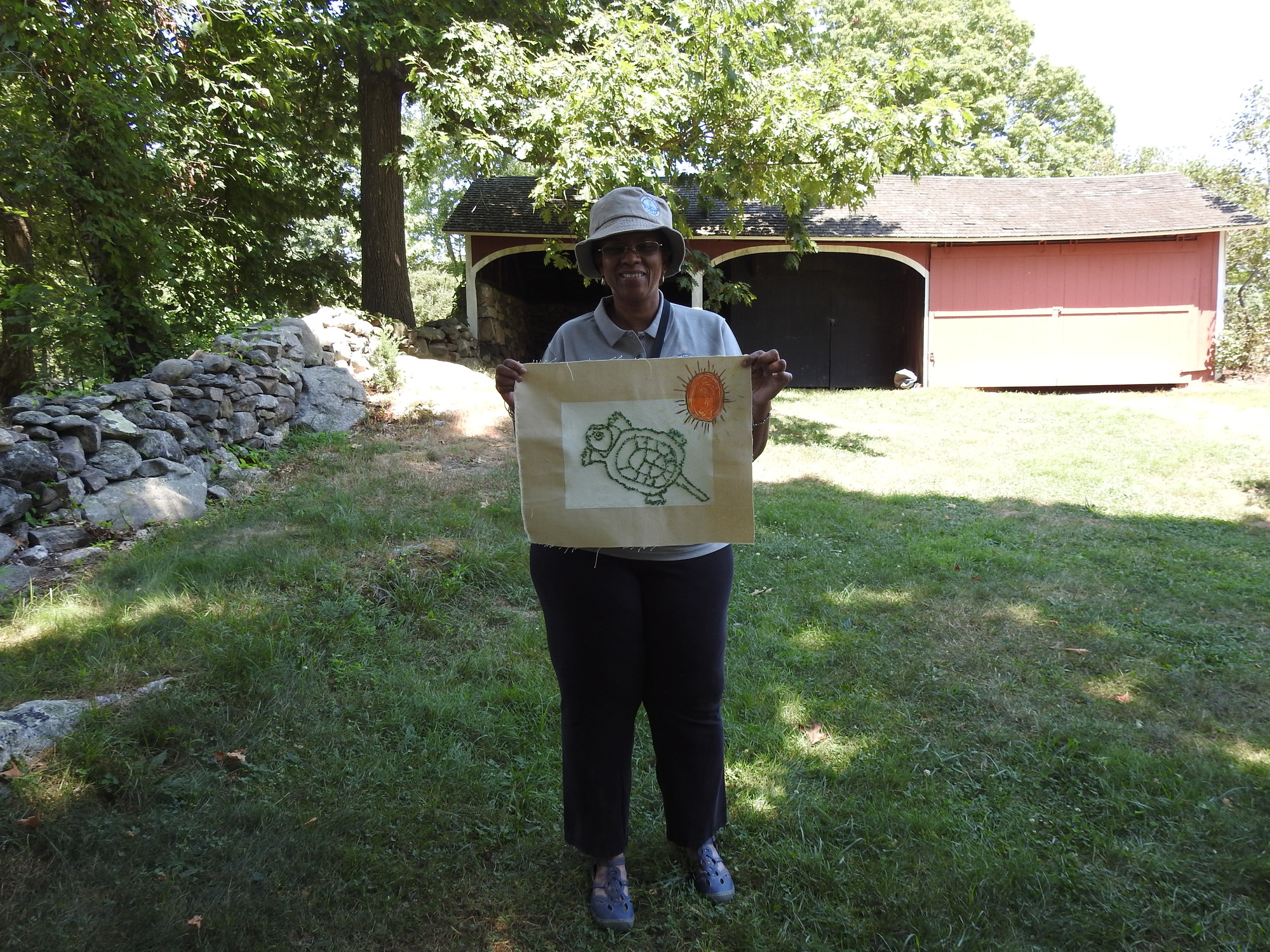 Weir Farm NHP Teacher-Ranger-Teacher holds up artwork of a turtle with sun. They stand in front of a stone wall and red farm building.  
