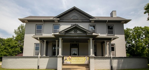 Two story brick home with multiple windows and front porch. Several trees with green leaves are in the background. Green banner with words and images of African American in military uniform.