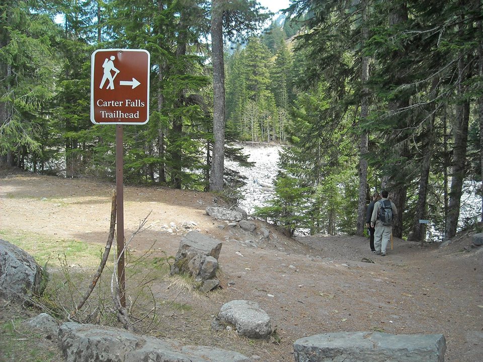 Hikers walk on a trail past a trail sign reading "Carter Falls Trailhead".