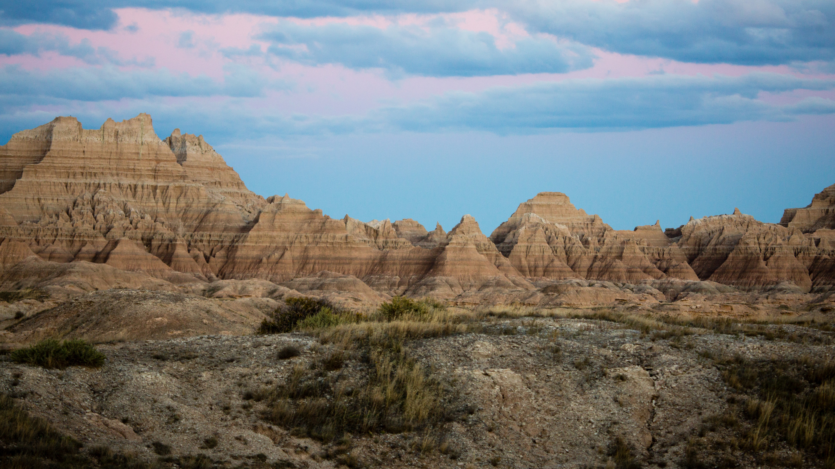 Several ridges and hills rise above the plains at Badlands National Park. The ridges and hills have many different layers that are made of different kinds of rock. Below the ridges and hills there are patches of grass and bare ground. 