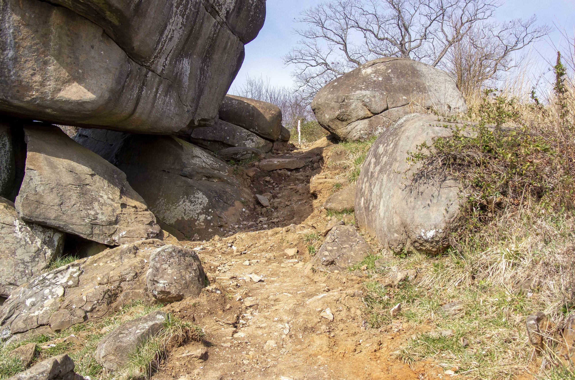 The camera is looking up a path along the slope of a small hill. The path is plain brown with its pavement recently removed. A massive boulder formation dominates the left portion of the frame. There are smaller boulders on the opposite end of the path.