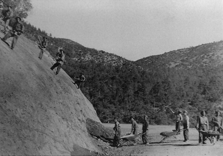Black and white photo of men working on a road--some are hooked into ropes on a steep rock face and others are holding shovels and wheelbarrows. 