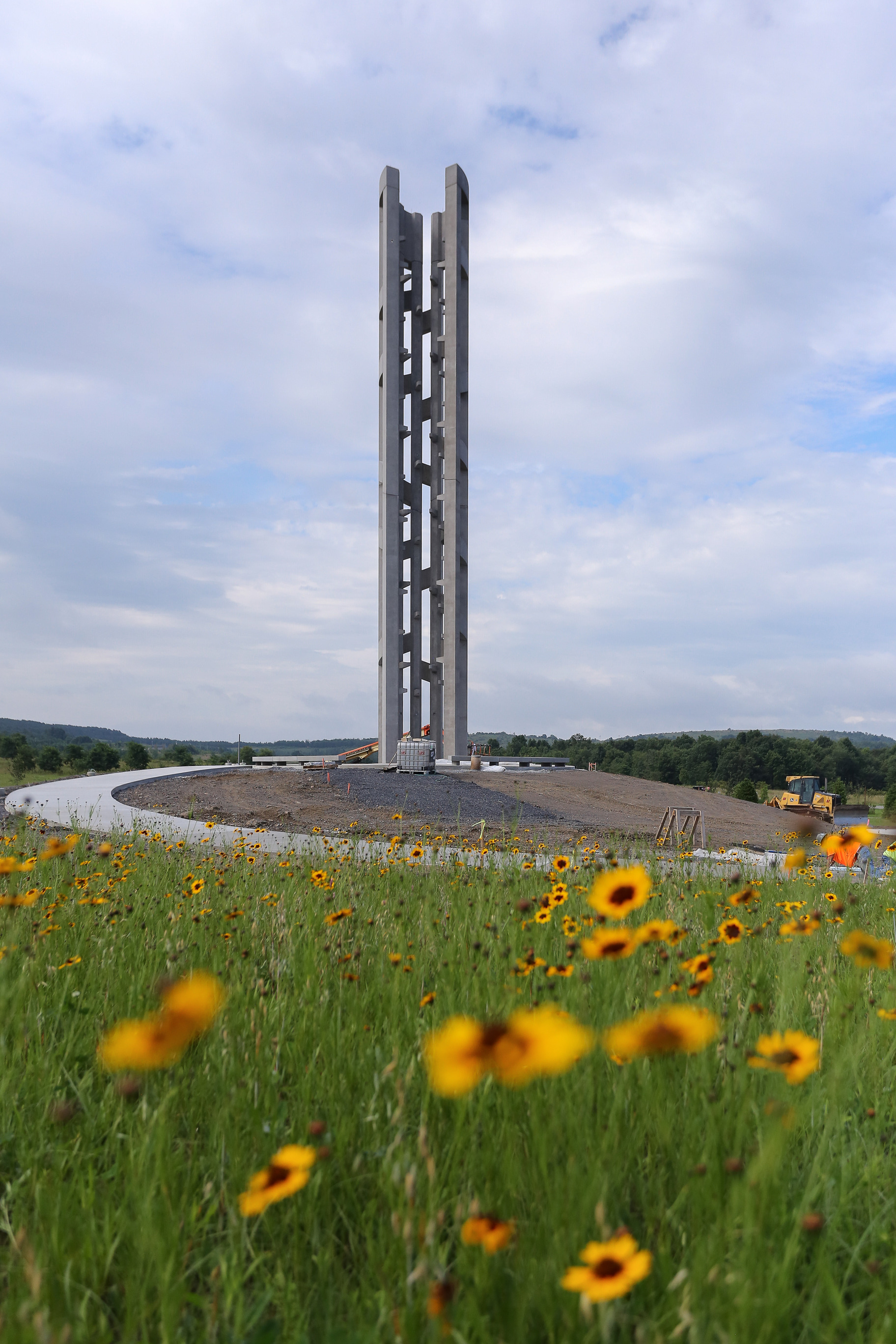 The Tower of Voices is in the center of grounds construction. A gentle rise of trees is in the distance and the green field in the foreground is dotted with small yellow wildflowers.