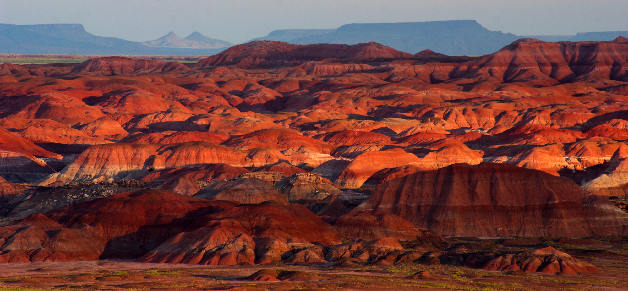 Red badlands in front of distant bluish mesas, mostly cloudy with a small sunny highlight.