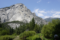 A river flows in the foreground in between grasses, bushes, and trees. Behind them is a large granite mountain dotted with trees. 