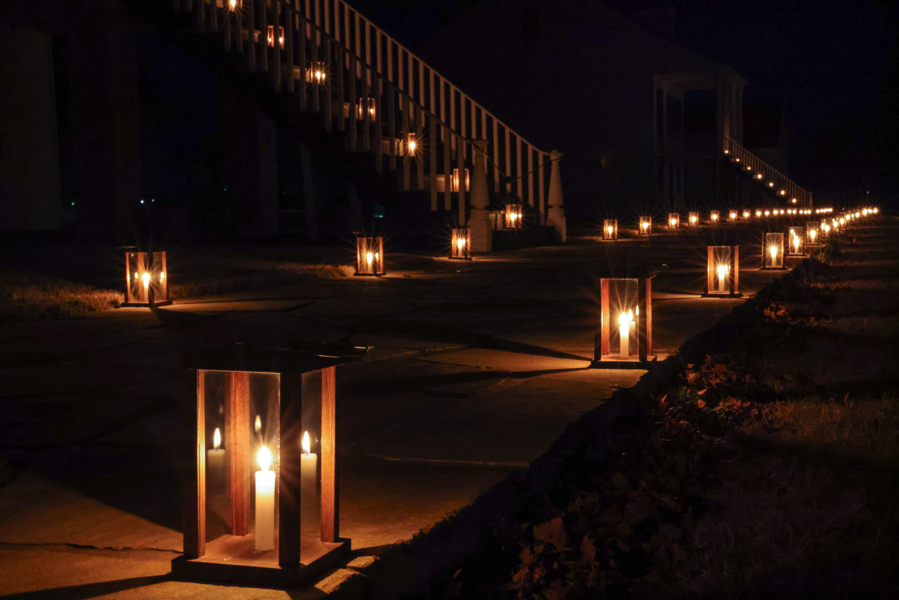 Candle lanterns line a walkway and go up the stairs of two staircases. Nearly everything not in close proximity to the lanterns is darkened or black.