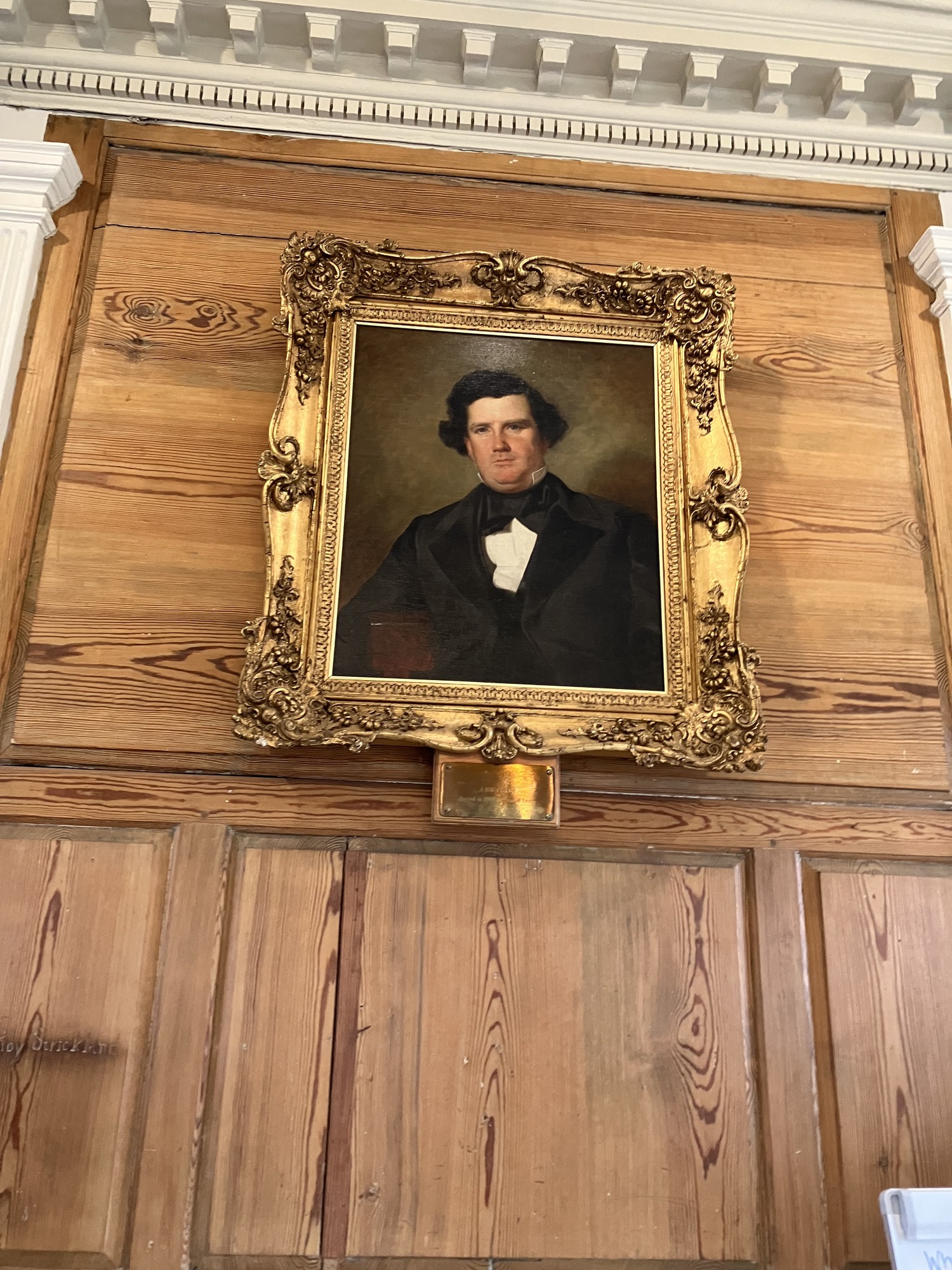 Against a bare wood-paneled wall with cream-painted trim hangs an oil painting of a middle-aged white man in a 19th Century-style suit, with an ornate gold frame around it.