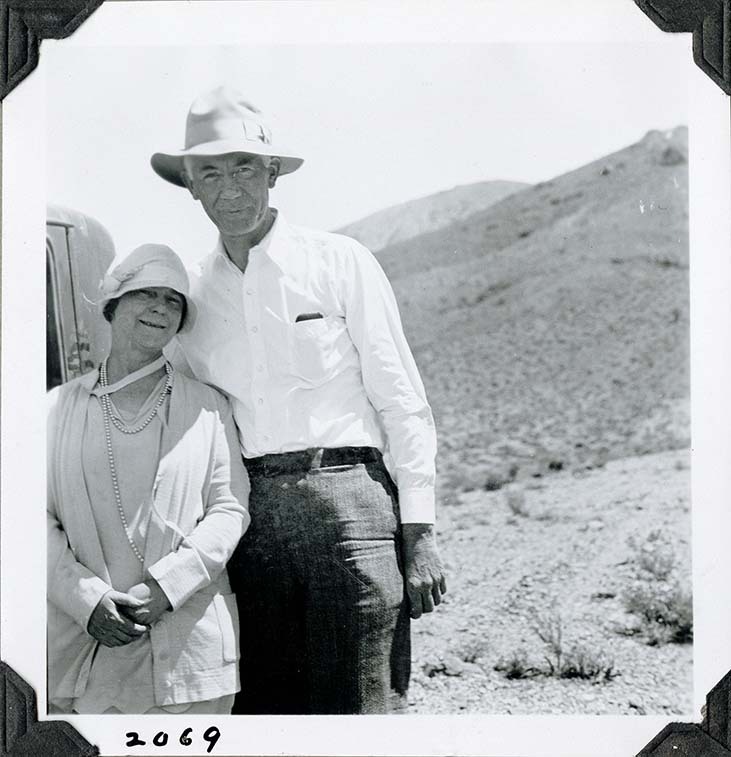 This is an historic black and white photograph from the Scotty's Castle Historic Photograph Collection, Death Valley National Park of a middle-aged couple posing for the camera. Woman on left, shorter, light colored 1920s dress and hat, leaning into man. Man on right, tall, floppy western hat, light colored shirt with something in front pocket, dark slacks. Cab of vehicle behind. Background of barren desert hillsides. Number in black ink in lower left corner.